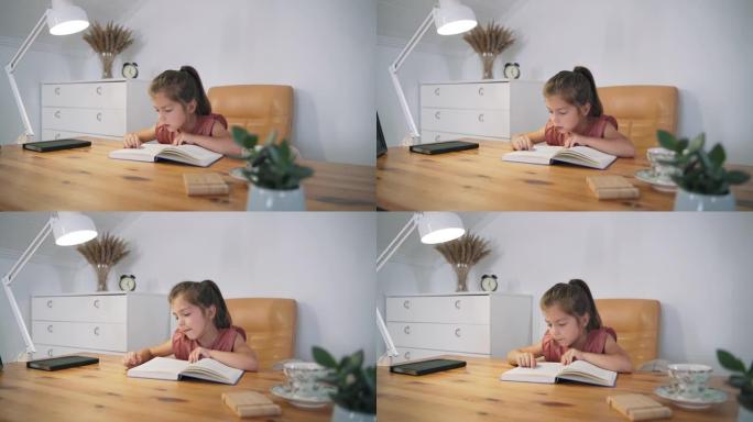 一个女孩坐在桌子旁看书。这孩子正在学习阅读。家庭教育。一个聪明的孩子。看书。儿童童话。自我隔离。慢动