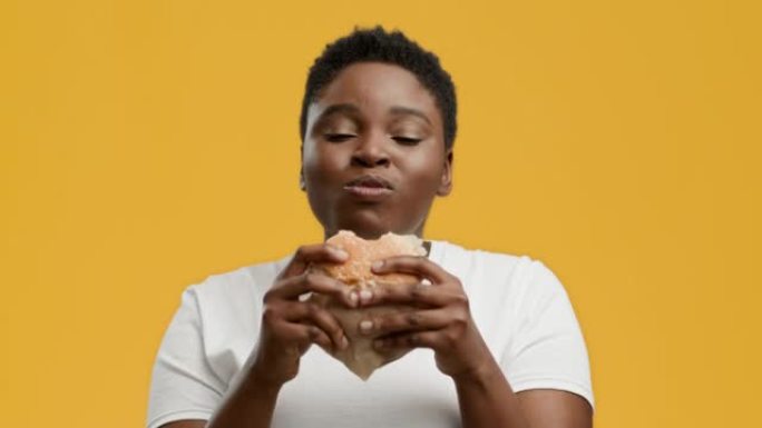 快乐肥胖的非洲裔美国妇女在黄色背景上吃汉堡
