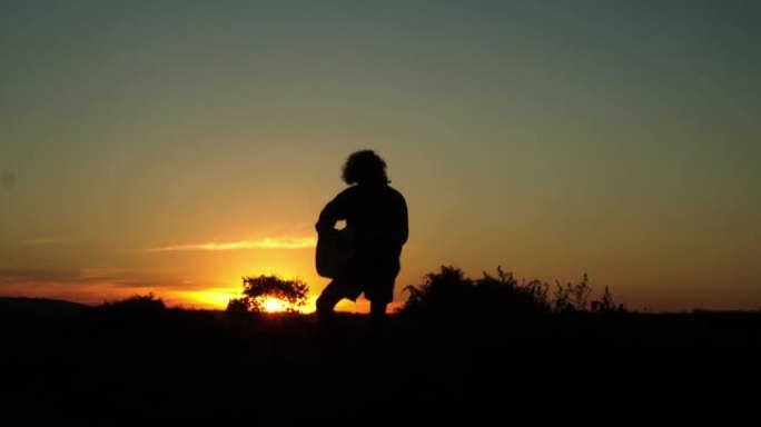 音乐家在日落时弹吉他的剪影，有日出或日落的音乐，大自然中的吉他，橙色的天空。4k慢动作