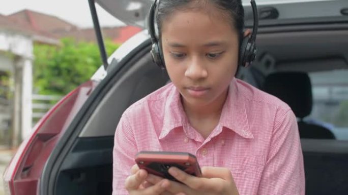 快乐可爱的亚洲少女戴着耳机，坐在住宅区掀背车的后侧，享受通过移动智能手机在线观看社交媒体的乐趣。