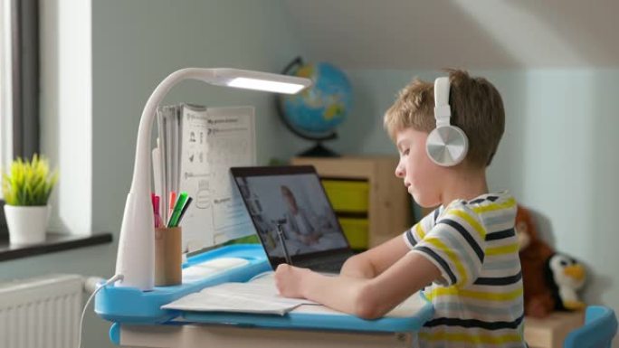 男孩坐在家里的笔记本电脑上。在线课程。男孩在网上问候老师。他挥手。