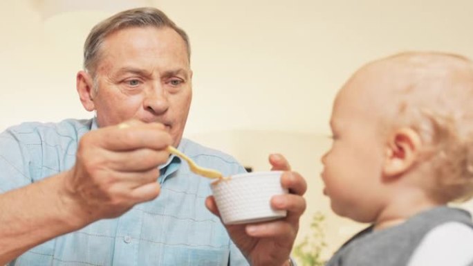 一个成熟的老年人坐在喂食椅上的桌子上喂孙子，蹒跚学步的孩子用碗中的勺子吃祖父提供的晚餐，孩子对世界感