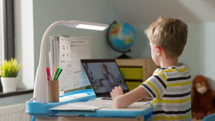 男孩坐在家里的笔记本电脑上。在线课程。孩子向老师打招呼。屏幕上的老师打招呼，手里拿着铅笔的男孩开始学
