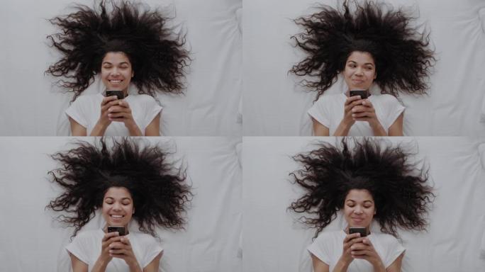 迷人的非洲裔美国女孩，卷发在白色的床上，使用智能手机，在社交媒体上写博客或与朋友聊天并微笑。顶视图
