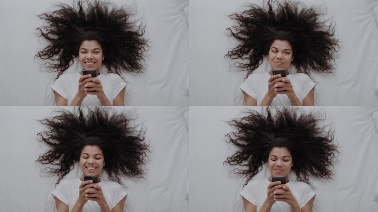 迷人的非洲裔美国女孩，卷发在白色的床上，使用智能手机，在社交媒体上写博客或与朋友聊天并微笑。顶视图