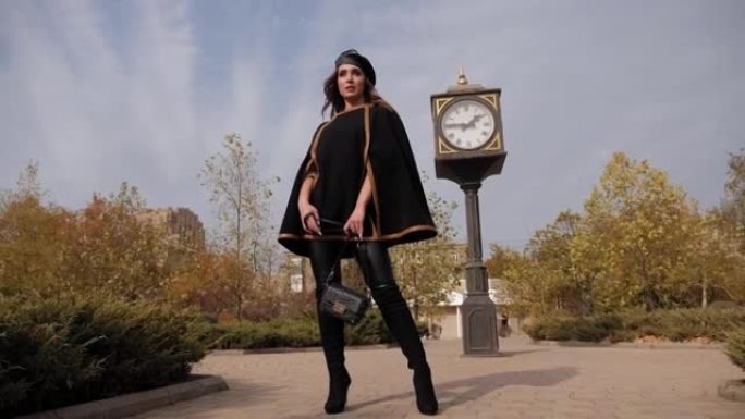 一位穿着黑色斗篷的年轻优雅女子穿过城市的秋季公园。