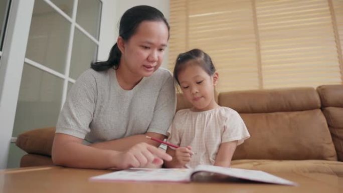 亚洲母亲照顾她的小女孩在家学习学校练习册上的写作