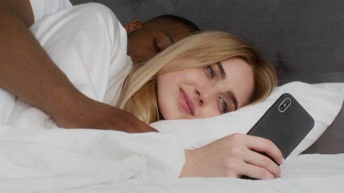 年轻的女士躺在床上，用智能手机联网，非洲裔美国丈夫睡觉并拥抱她