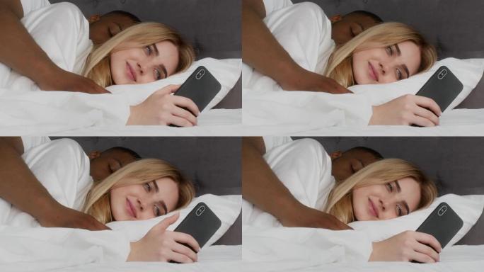年轻的女士躺在床上，用智能手机联网，非洲裔美国丈夫睡觉并拥抱她