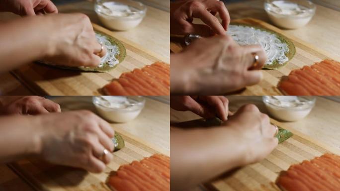 薄薄的美味彩色煎饼。女人的手放在鲑鱼片上的特写镜头，填充在木板上涂上奶油奶酪的绿色煎饼上。滚动煎饼。