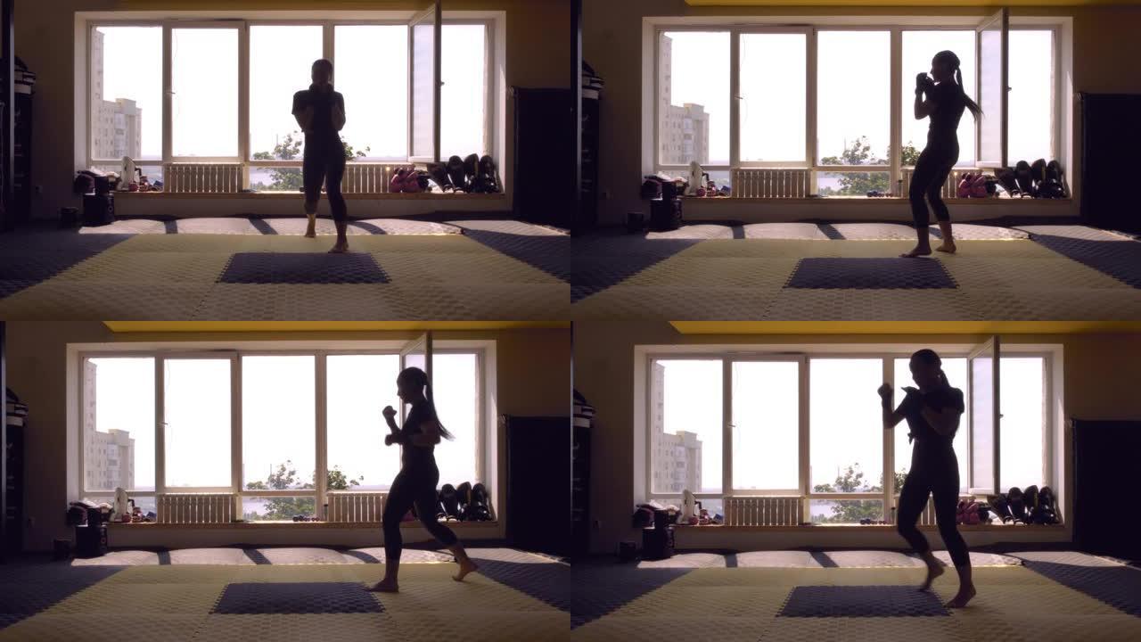 一个亚洲拳击手女孩的剪影在体育拳击馆一个大窗户的背景下。女孩罢工了。影子拳击