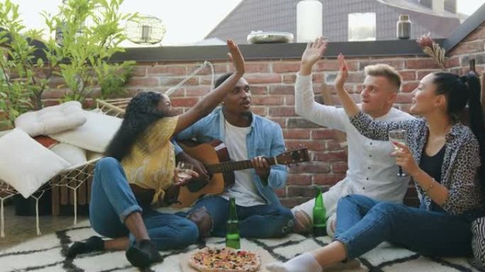 精神振奋的脾气暴躁的现代混合种族的前视图满足了年轻人，他们在夏季聚会期间在露台上享受着吉他，饮料和食