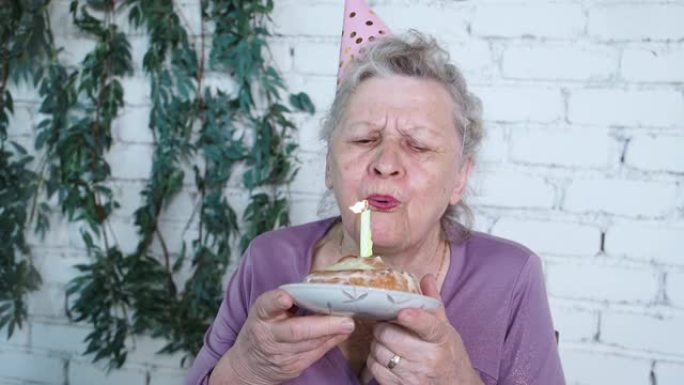 慢动作。快乐微笑的老奶奶庆祝85岁生日，拿着蛋糕吹出蜡烛感到感激，白砖背景。享受、尊重和温暖的关系。