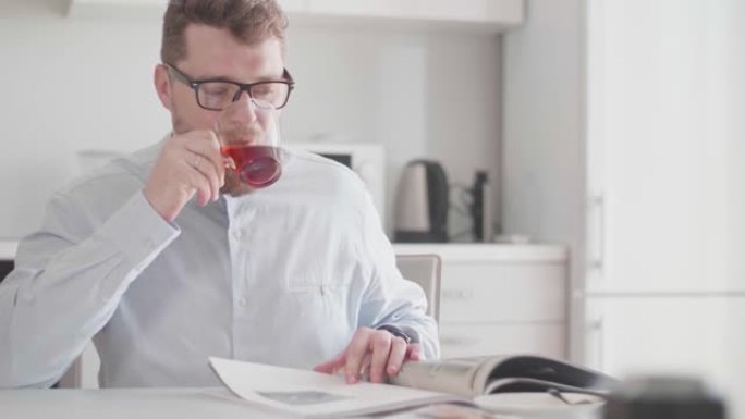 一个年轻的时尚男人早上在早餐时戴着眼镜看杂志并喝茶