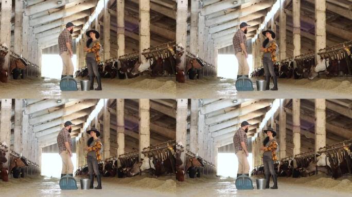 4k视频中年女农场工人戴着牛仔帽，拿着铲子，在牛棚里与年轻男工人讨论