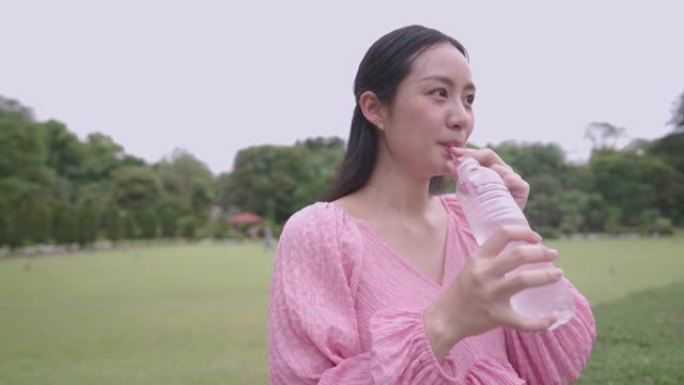 年轻的亚洲准妈妈喝水用吸管从塑料瓶中吸水，怀孕后期，孕妇喝水补液站在露天公园内的绿色草地上