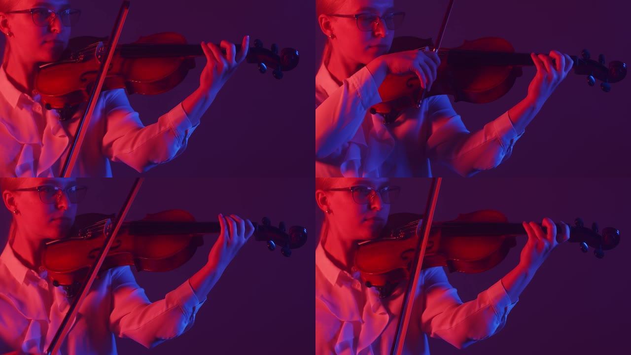 戴着眼镜和白色上衣的女小提琴手拉小提琴，站在工作室里，被红灯照亮。女音乐家用小提琴演奏古典音乐。交响