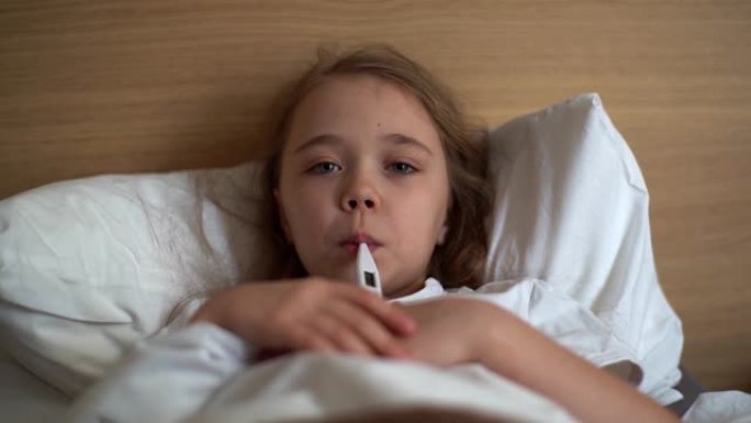发热高的生病小女孩的特写镜头用温度计测量自己的体温，躺在床上被毯子覆盖