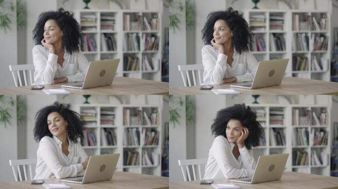 年轻的非洲裔美国妇女梦想着一些愉快和微笑的肖像。穿着白色上衣卷发的黑发女人坐在家庭办公室的桌子旁。特
