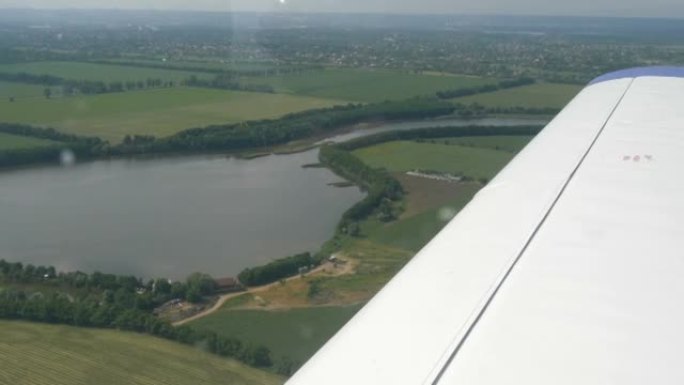 一架小型客机舷窗靠着白色机翼的视野。白色翅膀背景下的自然，河流和绿色田野的俯视图
