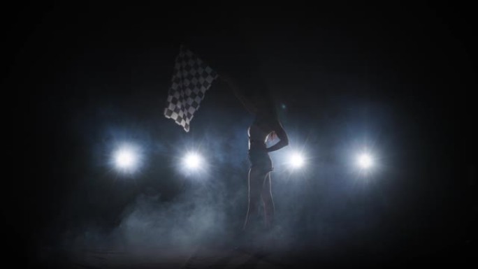 剪影的侧视图年轻女子挥舞着方格竞赛旗，标志着赛车活动的开始。黑发女人在带背光的黑暗烟熏工作室中摆出全