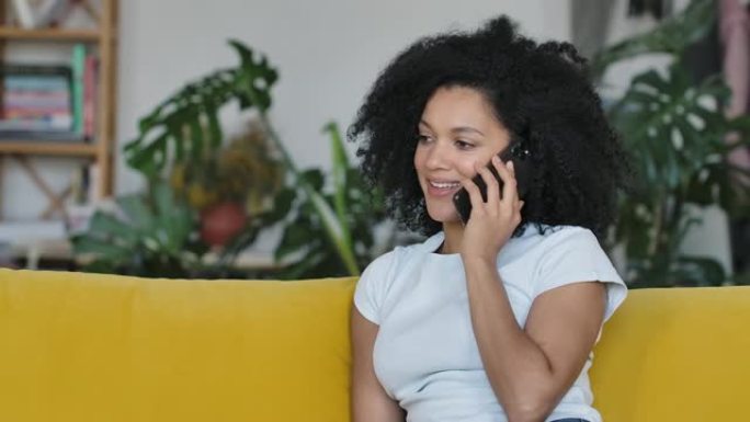 一位年轻的非洲裔美国妇女为手机聊天的肖像。黑发，卷发，坐在明亮的家庭房间里的黄色沙发上。特写。慢动作