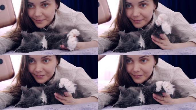 一个美丽的女人和一只灰猫躺在床上。女人举起宠物的爪子，为自己和宠物的照片拍了一张更有趣的照片。智能手