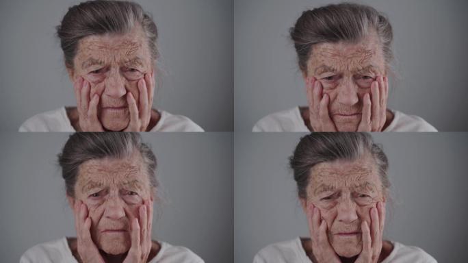 针对老年人的家庭暴力。受害人高级妇女害怕并哭泣，用手摩擦眼睛，看着灰色背景上的相机。成熟的女性双手在