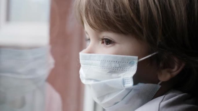 戴着防护面具的小男孩，悲伤地透过窗户看着担心新型冠状病毒肺炎封锁。戴着保护面具的孩子看着窗外的家。检