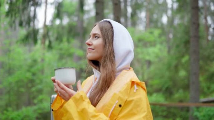 在潮湿的雨天，穿着黄色雨衣的女性旅行者从站在乡村木制门廊上的保温瓶中喝热茶。享受森林度假和白日梦的女