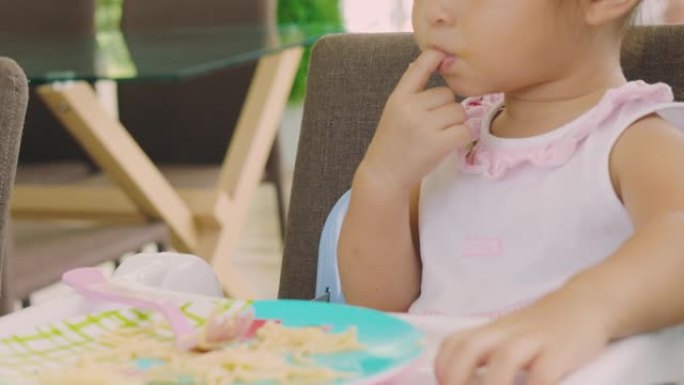 特写4k无法辨认的可爱的亚洲3岁宝宝正在吮吸手指，坐在椅子上用肮脏的手和嘴吃饭。她看起来饿了，喜欢吃