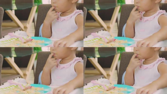 特写4k无法辨认的可爱的亚洲3岁宝宝正在吮吸手指，坐在椅子上用肮脏的手和嘴吃饭。她看起来饿了，喜欢吃