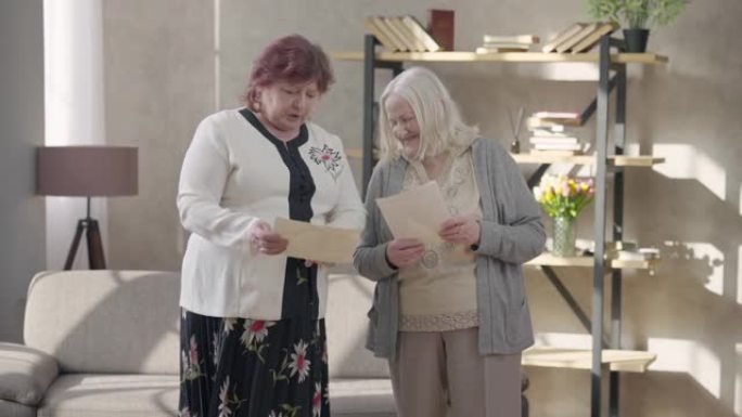 两名积极的高级退休人员笑着看着照片。女性高加索老朋友的肖像聊天回忆站在室内的客厅。友谊和怀旧理念