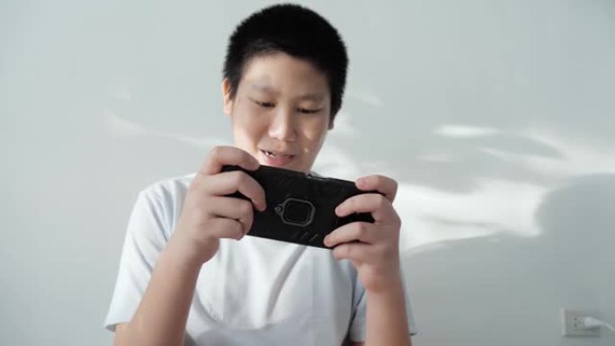 亚洲男孩坐在地板上玩手机游戏，家里有阳光阴影，生活方式概念。
