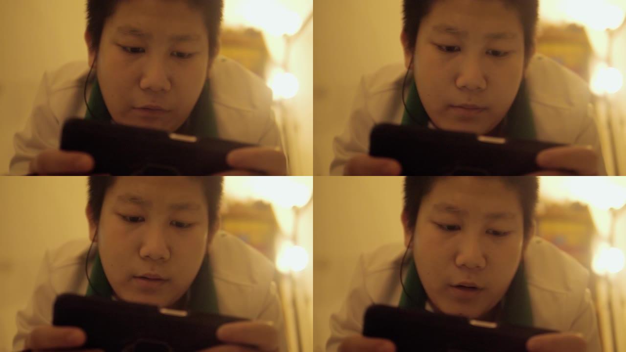 亚洲男孩在夜间通过智能手机在床上玩网络游戏，拥有温暖的光线，生活方式的概念。