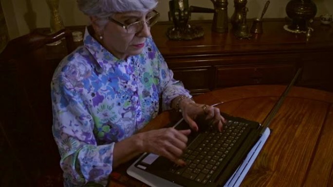 一位拉丁美洲高级女士在家中坐在餐桌旁的笔记本电脑上工作