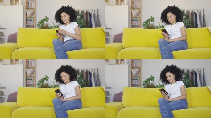 一位年轻的非洲裔美国妇女在手机上发短信的肖像。黑发，卷发，坐在明亮的家庭房间里的黄色沙发上。慢动作就