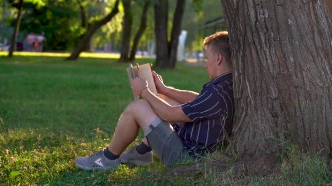 4k视频。一个年轻人坐在公园草地上的树下，在日落时看书。俄罗斯圣彼得堡