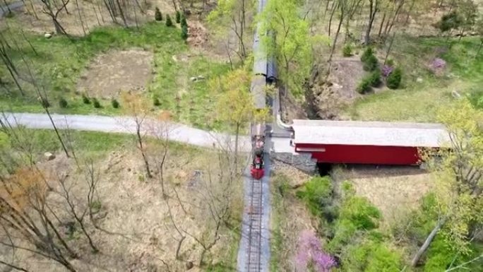 一列19世纪60年代蒸汽客运列车在一条孤独的单轨轨道上经过一座有盖的桥时，在林区行驶的鸟瞰图