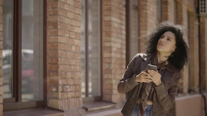 时尚的年轻非洲裔美国妇女的肖像在手机上看着地图找到了交汇点。穿着棕色皮夹克的黑发女人在街上摆姿势。特