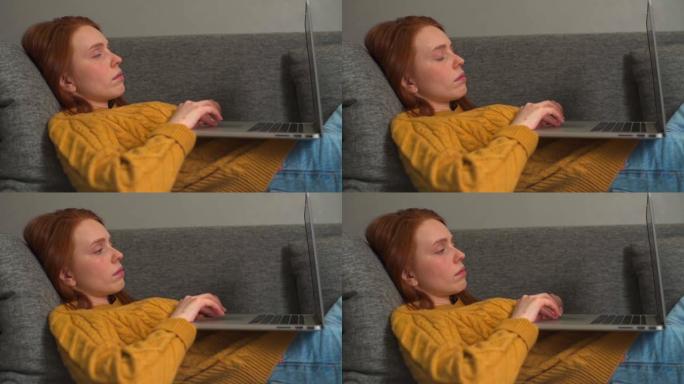 昏昏欲睡的红发年轻女子躺在家里沙发上时在笔记本电脑上打字的特写。