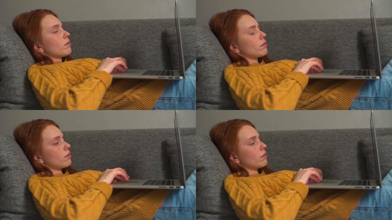 昏昏欲睡的红发年轻女子躺在家里沙发上时在笔记本电脑上打字的特写。