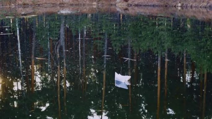 被风吹开的孤独纸船平静地漂浮在水面上。针叶树在水中反射。折纸船