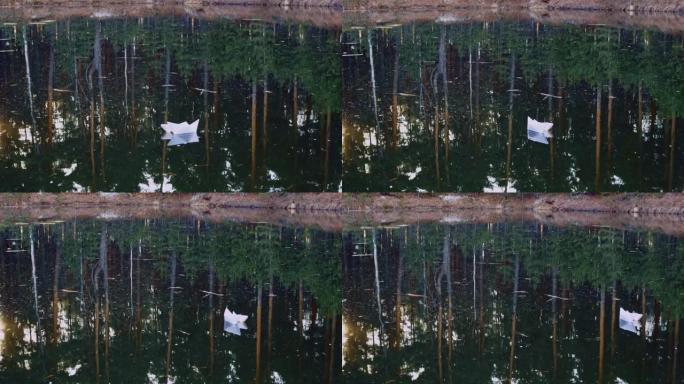 被风吹开的孤独纸船平静地漂浮在水面上。针叶树在水中反射。折纸船
