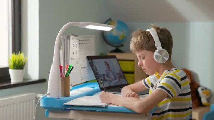 男孩坐在家里的笔记本电脑上。在线课程。戴着耳机的孩子听在线课程，然后在笔记本上写字。学生举手获得发言