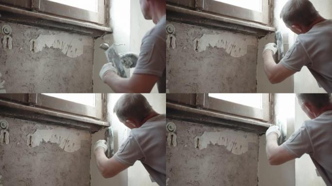 熟练的修理工在工作服上使用金属抹刀，刮刀和白色的抹灰糊将破旧的水泥墙放在窗下，用腻子粉刷平整墙面。墙