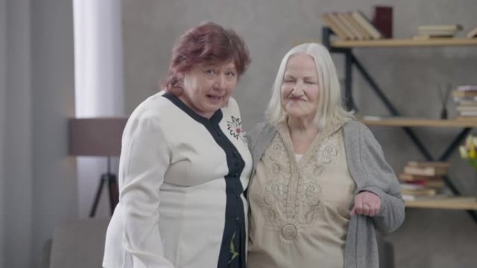 两个活跃的高级妇女在客厅里看着相机跳舞。积极的女性退休人员的肖像在周末在室内聚会很开心。快乐与退休生