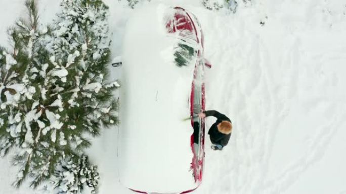 年轻女子早上在停车场自上而下的视野中清理一辆冰雪覆盖的汽车。美丽的女人在寒冷的冬天早晨刷她的雪车。