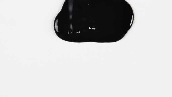 细流中的黑色油漆向下流到白色表面上，形成扩散的污渍并飞溅。特写白色背景上的黑色印迹。滴墨水。溢出油漆