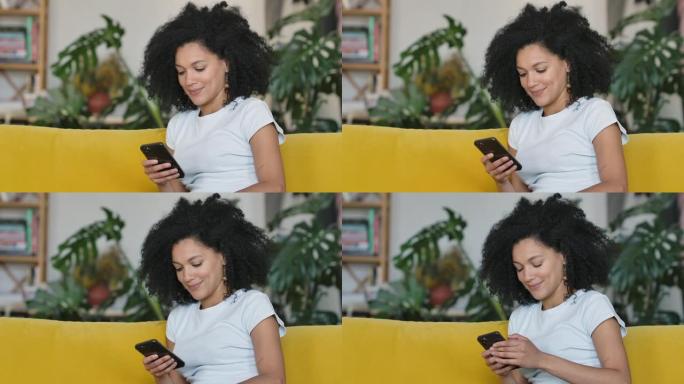 一位年轻的非洲裔美国妇女在手机上发短信的肖像。黑发，卷发，坐在明亮的家庭房间里的黄色沙发上。特写。慢